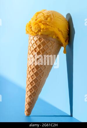 Eis in einem Waffelkegel auf einem blauen Tisch in hellem Licht. Mango-Eislöffel in einem Kegel, auf einem blauen Hintergrund angelehnt. Stockfoto