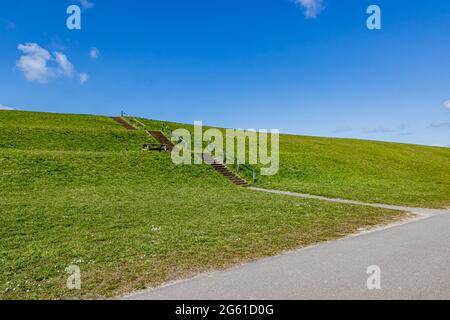 Deich am Strand von Camperduin mit grünem Gras und Betontreppen mit einem Metallgeländer, sonniger Frühlingstag mit einem blauen Himmel und einigen weißen Wolken in N Stockfoto