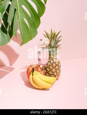 Frische Bananen, Ananas, Granatapfel und Erfrischungssaft im Glas auf pastellrosa Hintergrund. Natürlicher grüner Pflanzenschatten. Tropische Sommerszene. Stockfoto