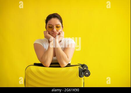 Unglückliche Frau in Sonnenbrille umarmt einen Koffer auf gelbem Hintergrund. Ein verärgertes Mädchen verpasste ihren Flug im Flugzeug. Annullierung aller Flüge aufgrund von Stockfoto