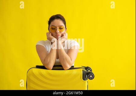 Unglückliche Frau in Sonnenbrille umarmt einen Koffer auf gelbem Hintergrund. Ein verärgertes Mädchen verpasste ihren Flug im Flugzeug. Annullierung aller Flüge aufgrund von Stockfoto