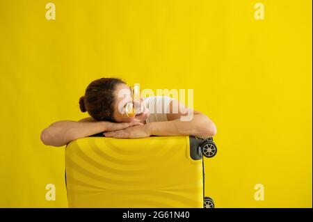 Lächelndes Mädchen in Sonnenbrille legte ihren Kopf auf einen Koffer auf gelbem Hintergrund. Glückliche Frau mit Handtasche wartet auf die Reise. Gepäck auf Rädern. Stockfoto