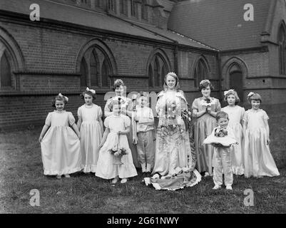 1956, historisch, draußen auf dem Gelände einer Kirche, eine May Queen, die in ihrem langen Kleid für ein Foto steht und einen Blumenstrauß mit ihrem Gefolge von Helfern, jungen Mädchen und zwei kleinen Jungen hält, von denen einer ein Kissen mit ihrer Krone in der Hand hält. Der Maifeiertag, ein Fest uralter Herkunft, das zur Feier der Ankunft des Sommers in Dörfern in ganz Großbritannien abgehalten wurde, mit der Krönung der May Queen, eines Mädchens und einer Parade, bei der sie der lokalen Bevölkerung ihr Kleid und ihre Krone zeigt. Stockfoto