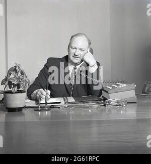 Der historische Manager aus den 1970er Jahren sitzt mit Pad und Stift am Schreibtisch und spricht am Telefon, England, Großbritannien, mit der Obilgatory Indoor House plant und dem Glasaschenbecher auf dem Schreibtisch. Stockfoto