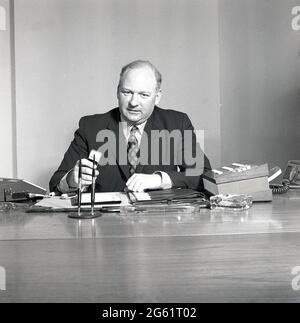 70er Jahre, historische, Exekutive sitzt an seinem Schreibtisch und spricht in die ?? Einer Diktatmaschine oder einem Sprachaufnahmelaufwerk, England, Großbritannien, mit dem Obilgatory Indoor House plant und dem Glasaschenbecher auf dem Schreibtisch. Stockfoto