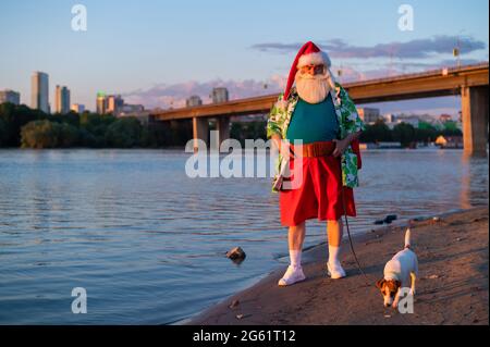 Der Weihnachtsmann in Shorts geht mit dem Hund Jack Russell Terrier am Strand entlang. Stockfoto