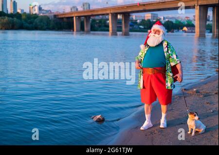 Der Weihnachtsmann in Shorts geht mit dem Hund Jack Russell Terrier am Strand entlang. Stockfoto