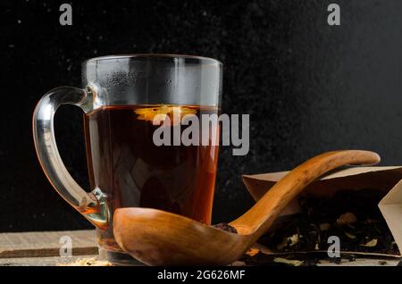 Ein Glas mit duftendem, gebrühtem Blumentee auf einem Holztisch mit einem Holzlöffel. Schwarzer Hintergrund. Selektiver Fokus. Stockfoto