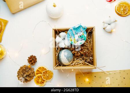 Weihnachts-Geschenkbox mit Souvenir aus Fimo. Zapfen, getrocknete Orangen, Girlande und Bastelpapier auf weißem Tisch. Neujahrsstimmung. Tolle Komposition für die Gestaltung von Grußkarten. Stockfoto