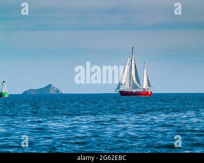 Yacht im klassischen Stil mit rotem Rumpf segelt an grüner Boje vorbei auf dem wunderschönen tiefblauen Meer. Mit entfernter Insel. Stockfoto