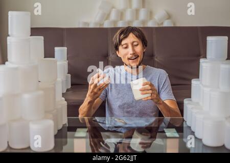 Der Mann isst Joghurt und ist von vielen Joghurtgläsern umgeben, die er in einem Jahr verwendet. Eine riesige Menge von Plastikbehältern für Lebensmittel. Übermäßiger Verbrauch von Stockfoto