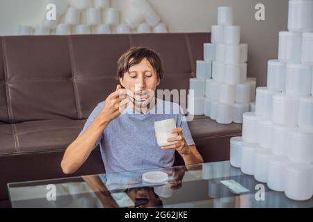 Der Mann isst Joghurt und ist von vielen Joghurtgläsern umgeben, die er in einem Jahr verwendet. Eine riesige Menge von Plastikbehältern für Lebensmittel. Übermäßiger Verbrauch von Stockfoto