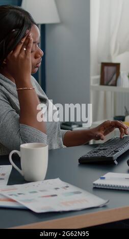 Müde verzweifelte schwarze Student leiden Kopfschmerzen sitzen am Schreibtisch im Wohnzimmer auf der Suche nach medizinischen Behandlung mit Computer. Besorgt krank verärgert junge Frau mit Migräneschmerzen während Lockdown Stockfoto