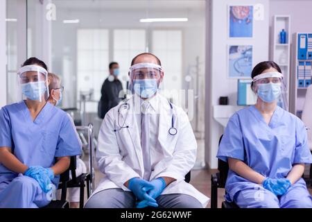 Eine Gruppe von Krankenschwestern und einem Arzt in Laborkittel und -Peelings, die die Kamera mit Gesichtsmaske und Visier gegen Coronavirus im Empfangsbereich des Krankenhauses betrachten. Patienten, die soziale Distanz respektieren Stockfoto