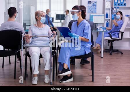 Medizinisches Personal mit Visier gegen Coronavirus im Krankenhaus im Gespräch mit behinderter alter Frau im Rollstuhl Ausfüllen des Formulars vor der Konsultation. Patienten, die während einer globalen Pandemie von Covid 19 einen Arzt besuchen Stockfoto