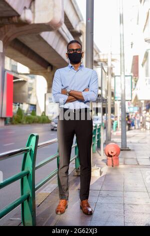 In voller Länge Aufnahme von schönen schwarzen afrikanischen Geschäftsmann im Freien in der Stadt im Sommer trägt Gesichtsmaske vor Corona-Virus zu schützen Stockfoto