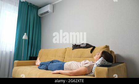 Die Frau schaltet die Klimaanlage über die Fernbedienung ein und liegt mit einer Katze auf der Couch. Stockfoto