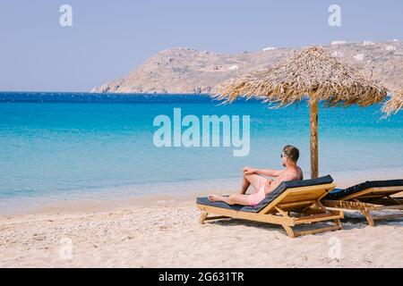 Elia Beach Mykonos, ein junger Mann am Strand von Mykonos im Sommer mit einem Sonnenschirm und luxuriösen Liegen, das blaue Meer am Elia Beach Mykonos Griechenland. Stockfoto