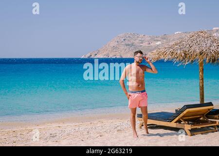 Elia Beach Mykonos, ein junger Mann am Strand von Mykonos im Sommer mit einem Sonnenschirm und luxuriösen Liegen, das blaue Meer am Elia Beach Mykonos Griechenland. Stockfoto