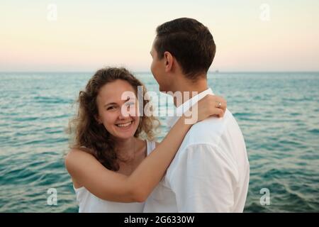 Frau umarmt ihren geliebten Mann und lächelt gegen den Sonnenuntergang am Meer Stockfoto