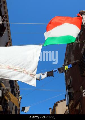 In der engen Straße der typisch mediterranen Stadt hängen viele Kleider, die in der Sonne trocknen, mit einer italienischen Flagge Stockfoto