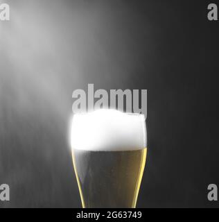 Ein Glas schaumiges kaltes Bier mit Rauchaerosol, Wasserstaub spritzt über, vor schwarzem dunklen Hintergrund. Stockfoto