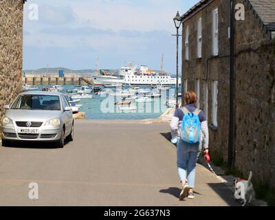 Eine Frau geht mit ihrem Hund in Richtung Hafen von Hugh Town, mit dem Scillonian III, der im Hintergrund vertäut ist, St. Mary's, Isles of Scilly, Cornwall, Großbritannien Stockfoto