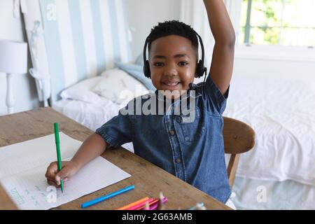 Lächelnder afroamerikanischer Junge, der zu Hause einen Videoanruf abhält und Kopfhörer trägt und die Hand hebt Stockfoto