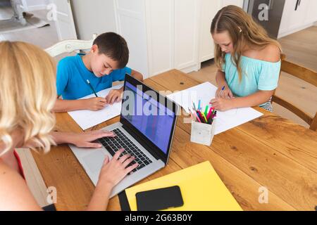 Kaukasische Mutter mit Laptop und ihre Tochter und ihr Sohn machen Hausaufgaben in der Küche Stockfoto