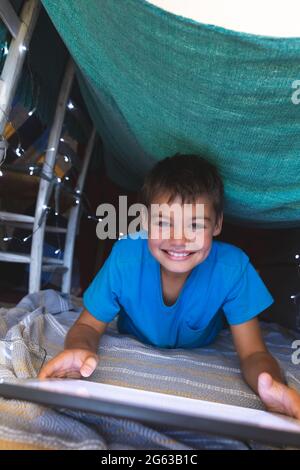 Porträt eines lächelnden kaukasischen Jungen, der zu Hause mit einem Tablet auf einer Decke in Fort liegt Stockfoto