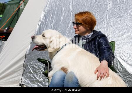 Russland. Vyborg 06.05.2021 EINE Frau sitzt mit einem weißen Labrador auf den Knien in der Nähe des Zeltes. Ruhen Sie sich mit Tieren in einer Spinne aus. Hochwertige Fotos Stockfoto