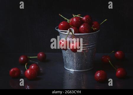 Rote Kirschen in einem eisernen Eimer auf schwarzem Hintergrund. Reife saftige Kirschbeeren in Wassertropfen auf dem Tisch. Das Konzept der Frische, richtige Nutri Stockfoto