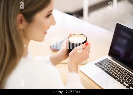 Schöne kaukasische Frau, die an etwas denkt, während sie mit einem Notebook in einer minimalistischen Café-Bar sitzt. Charmante Freiberuflerin, die während der Arbeit am Laptop beim Kaffeetrinken über neue Ideen nachdenkt Stockfoto