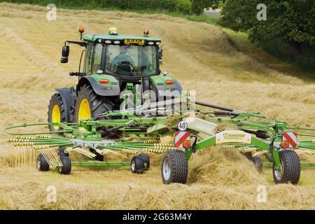 John Deere-Traktor, der während einer Heuernte auf einem Feld mit einem Doppelrotor-Heuwender oder Zetteraufsatz arbeitet Stockfoto