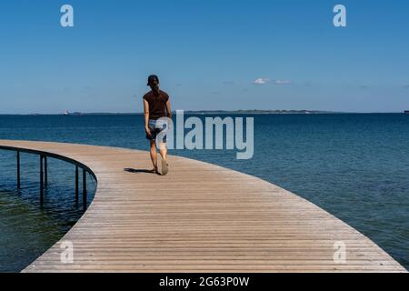 Eine schlanke Frau geht auf einer kreisförmigen Promenade entlang, die aus dem ruhigen, blauen Meer führt Stockfoto