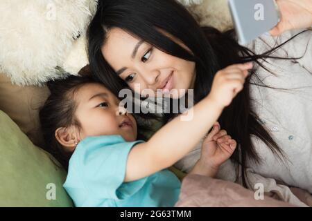 Asiatisches Mädchen zeigt auf Smartphone in der Nähe der Mutter und weiches Spielzeug auf dem Bett Stockfoto