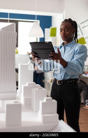 Architektin Frau afroamerikanischer Ethnie, die an Tablets arbeitet und ein professionelles Maquette-Baumodell ansieht. Architekturmitarbeiter betrachten Design für modernes Projekt in Entwicklung Stockfoto