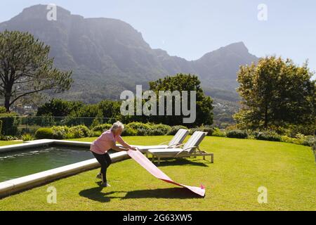 Ältere kaukasische Frau, die im sonnigen Garten eine Yogamatte ausrollt Stockfoto