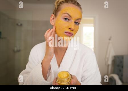Kaukasische Frau im Badezimmer trägt Bademantel, Blick in Spiegel und Anwendung Gesichtsmaske Stockfoto
