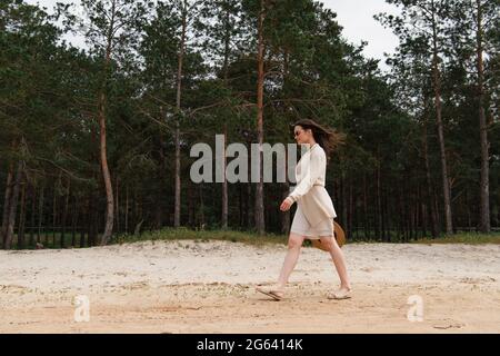 Die ganze Länge der fröhlichen jungen Frau in Sonnenbrillen mit Strohhut und Spaziergang in der Nähe des Waldes Stockfoto