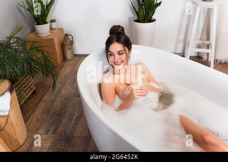 Man hat einen schönen Blick auf die glückliche Frau, die ein Glas Champagner in der Badewanne hält Stockfoto