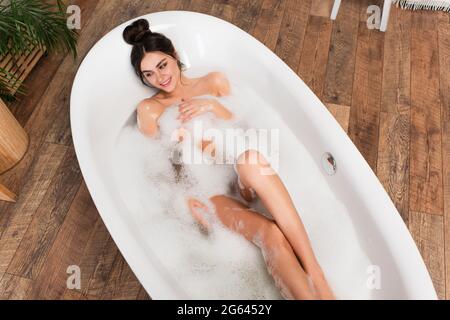 Blick von oben auf die junge fröhliche Frau, die in der weißen Badewanne baden geht Stockfoto
