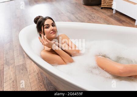 Aus dem hohen Winkel betrachtet sieht man eine fröhliche Frau mit einem Haarknoten, der die Kamera anschaut, während man in der Badewanne baden geht Stockfoto