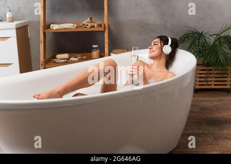 Eine Frau mit Kopfhörern, die beim Baden Musik hört und ein Glas Champagner in der Hand hält Stockfoto