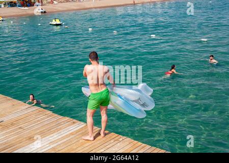 Antalya, Türkei-28. Juni 2021: Mann mit aufblasbarer Luftmatratze auf Holzdock, während andere Strandbesucher im Sommer in Antalya im Meer schwimmen. Stockfoto