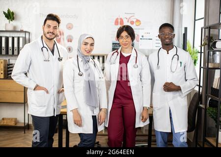 Porträt von vier multikulturellen Ärzten in weißen Laborkittel und Stethoskopen am Hals, die im Krankenhauszimmer posieren. Konzept der Medizin, Zusammenarbeit und Gesundheitsversorgung. Stockfoto