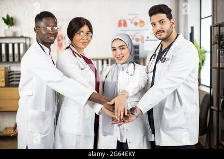 Eine Gruppe von vier multikulturellen Ärzten in weißen Laborkittel, die ihre Hände zusammenstapeln, während sie im Krankenhauszimmer von brigham stehen. Erfolgreiche internationale Zusammenarbeit. Stockfoto