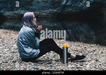 Teenager-Wandererin, die sich mit einem Becher am Flussufer ausruhte und auf die Küstenfelsen schaute Stockfoto