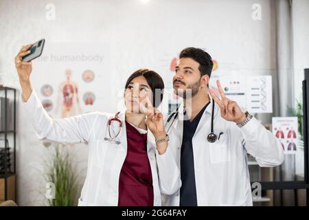 Lächelnde afrikanische Frau und indischer Mann im weißen Labormantel zeigen Zeichen Frieden mit den Fingern, während Selfie auf dem Smartphone. Zwei medizinische Mitarbeiter, die nach einem internationalen Treffen Fotos machen. Stockfoto