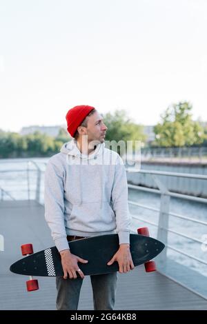 Junger Hipster Mann entspannt Skateboarder halten Longboard auf Brücke bei Sonnenuntergang. Urbanes Lifestyle-Konzept Stockfoto